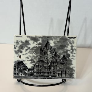 May include: A rectangular ceramic tile featuring a black and white transfer print of Trinity Church Boston. The tile includes the text Wedgwood, Copyright 1905 J.H.D. & S., and Trinity Church Boston. It is displayed on a black metal wire stand.