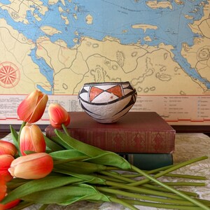 May include: A small, rounded ceramic bowl featuring a white base with black geometric line patterns and two orange triangular accents. The bowl is positioned on top of a stack of two books, surrounded by several orange and yellow artificial tulips.