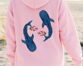 May include: A light pink hoodie featuring a blue whale shark design on the back. The whale sharks have white spots and are facing each other, with coral in between. The hoodie is a long sleeve with a hood.