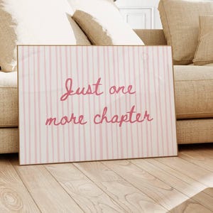 May include: A framed print with a gold border featuring the text "Just one more chapter" in pink script. The background has vertical pink and white stripes. The print is leaning against a beige sofa on a light wood floor.