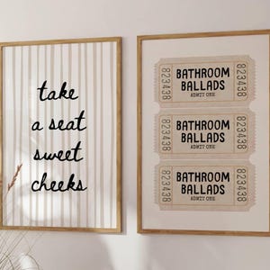May include: Two framed wall art pieces. One features vertical beige stripes with the text "take a seat sweet cheeks" in black. The other displays three vintage-style tickets with "Bathroom Ballads" and "Admit One".