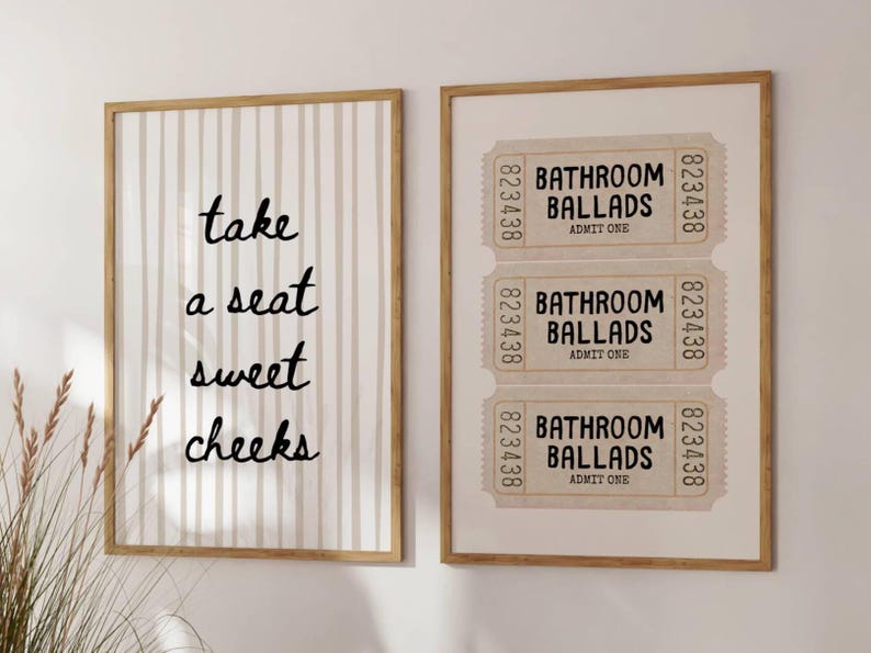 May include: Two framed wall art pieces. One features vertical beige stripes with the text "take a seat sweet cheeks" in black. The other displays three vintage-style tickets with "Bathroom Ballads" and "Admit One".