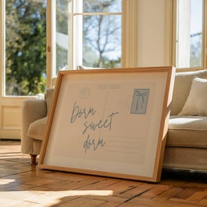 Pu&ograve; includere: Una stampa incorniciata di una cartolina vintage con il testo "Born sweet dorm" in un carattere blu chiaro. La cartolina ha un francobollo blu con un fiocco e uno sfondo marrone chiaro.