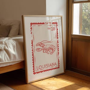 May include: A framed wall art print featuring a red graphic illustration of a lobster centered within a rectangular postage stamp design. The text USA 18 is printed at the top right, and the word LOUISIANA is printed at the bottom.