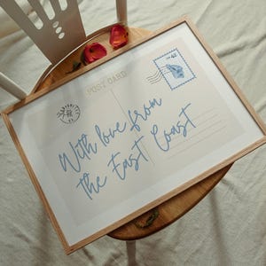 May include: A framed postcard print with the text "With love from the East Coast" in light blue script. The print has a vintage stamp and is displayed on a wooden surface.