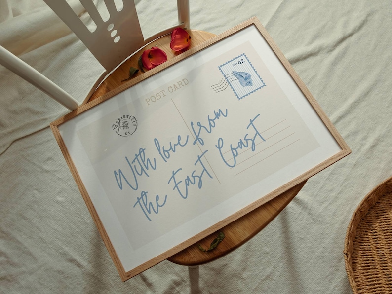 May include: A framed postcard print with the text "With love from the East Coast" in light blue script. The print has a vintage stamp and is displayed on a wooden surface.