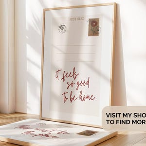 Pu&ograve; includere: Una stampa incorniciata con un design di cartolina vintage che presenta il testo "It feels so good to be home". La stampa &egrave; in una cornice di legno chiaro e ha uno sfondo bianco con testo rosso.