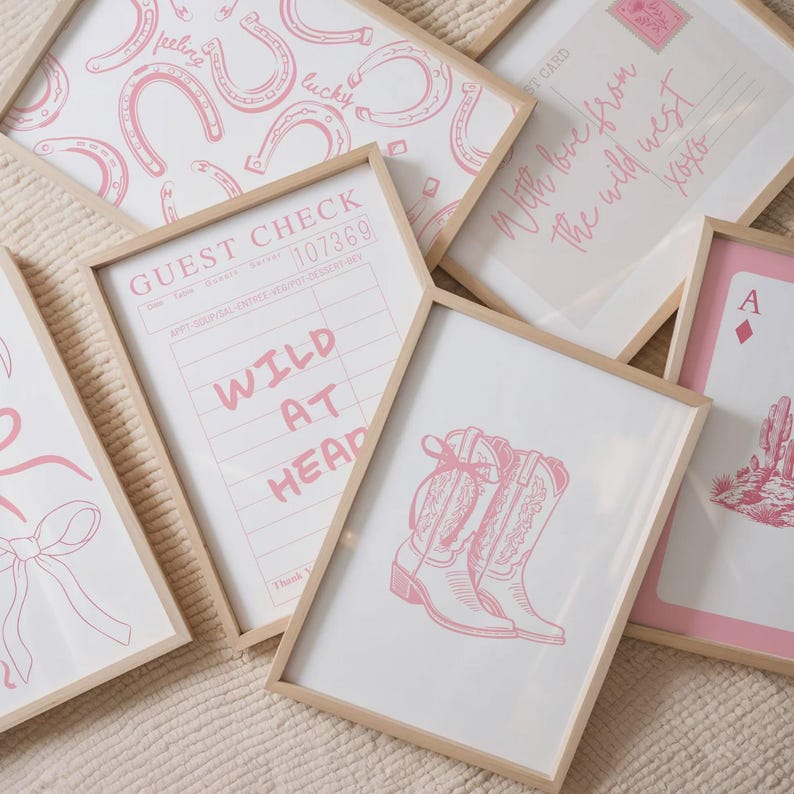 May include: A collection of framed wall art prints featuring pink illustrations on white backgrounds. Designs include a guest check with the text WILD AT HEART, cowboy boots, a horseshoe pattern, a postcard with text, and an ace of diamonds card.