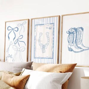 May include: Three framed art prints with a country theme. The prints feature blue bow illustrations, a horseshoe with a bow, and a pair of cowboy boots. The artwork is set against a white background with light brown wooden frames.