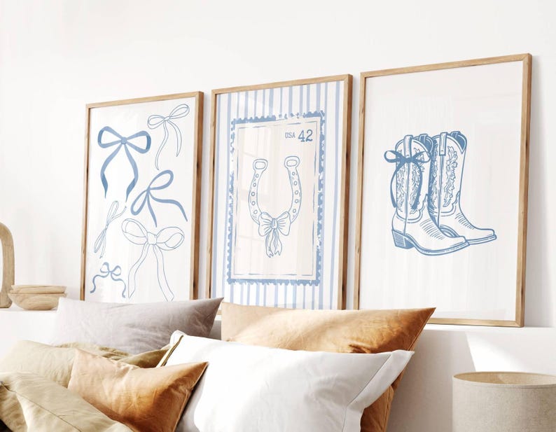May include: Three framed art prints with a country theme. The prints feature blue bow illustrations, a horseshoe with a bow, and a pair of cowboy boots. The artwork is set against a white background with light brown wooden frames.