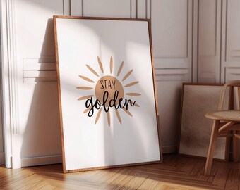 May include: A framed art print with a white background and a sunburst design in shades of tan and brown. The words "STAY golden" are written in black script. The frame is a dark brown color. The print is leaning against a wall.