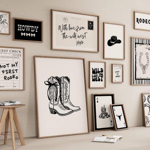 May include: A gallery wall display featuring multiple framed black and white prints. The artwork includes illustrations of cowboy boots, a horseshoe, a cactus, a bull skull, a cowboy hat, and text designs reading Howdy, Not My First Rodeo, and Wild Like The West.