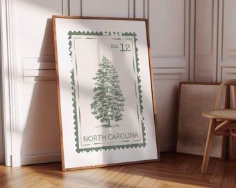 May include: A framed print with a green tree illustration, designed like a postage stamp. The text includes "USA 12" and "NORTH CAROLINA". The frame is dark brown. The print is leaning against a white wall, with a wooden floor in the foreground.