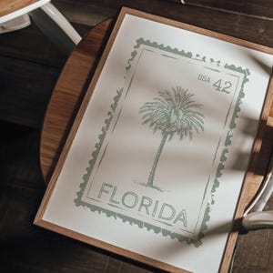 May include: A framed print featuring a palm tree design within a postage stamp border. The print is in shades of green and white, with the word "FLORIDA" and "USA 42" visible. The frame is made of wood.