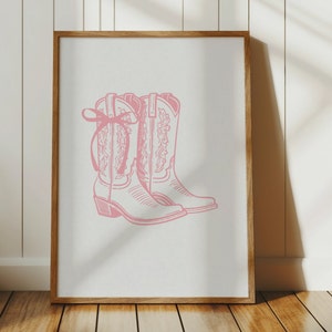 Op de afbeelding: Een ingelijst afdruk van een paar roze cowboylaarzen met een strik op een laars.