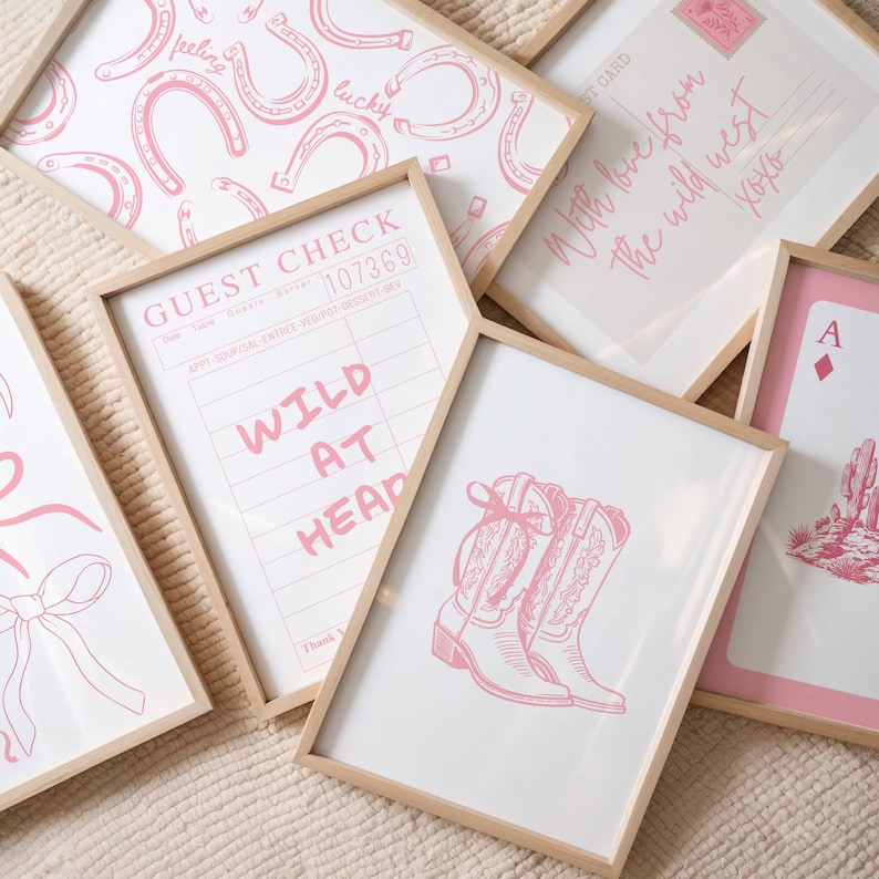 May include: A collection of framed art prints with a pink and white color scheme. The prints feature various designs, including horseshoes, cowboy boots, a guest check with "WILD AT HEAD" text, a postcard with text, a bow, and a playing card.