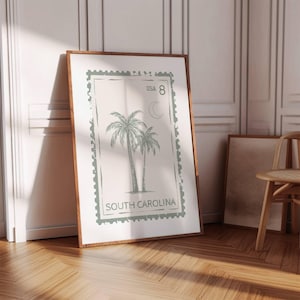 May include: A framed print with a vintage design of two palm trees, a crescent moon, and the text "SOUTH CAROLINA" and "USA 8". The artwork has a light green and white colour scheme, with a wooden frame.