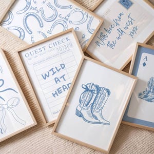 May include: A collection of framed art prints with a Western theme. The prints feature blue illustrations on white backgrounds, including horseshoes, cowboy boots, a guest check with "WILD AT HEAD", a postcard with "With love from the wild west xoxo", and a playing card.