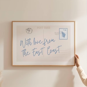 May include: A framed print resembling a vintage postcard. The design features the text "With love from the East Coast" in blue script, along with a postage stamp and postmark. The frame is a light brown color.