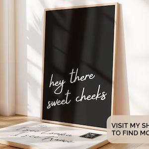 May include: A framed black print with the words "hey there sweet cheeks" in white script. A second print with text is visible in the foreground. The frame is light-colored wood. The image is in a well-lit room.