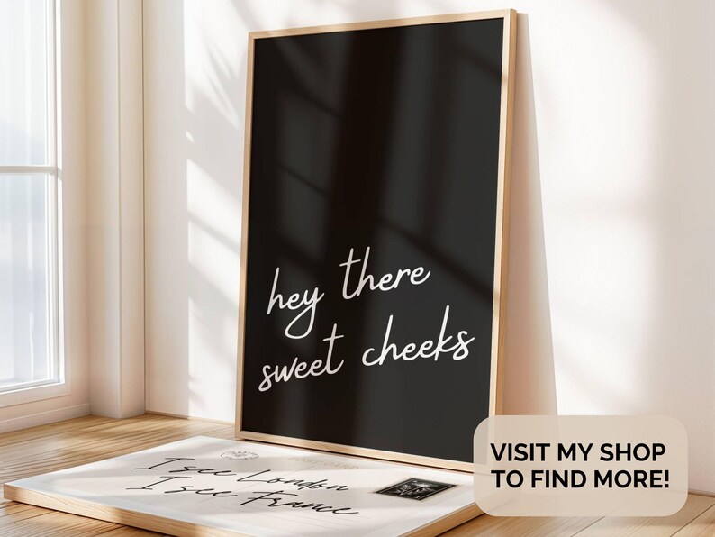 May include: A framed black print with the words "hey there sweet cheeks" in white script. A second print with text is visible in the foreground. The frame is light-colored wood. The image is in a well-lit room.