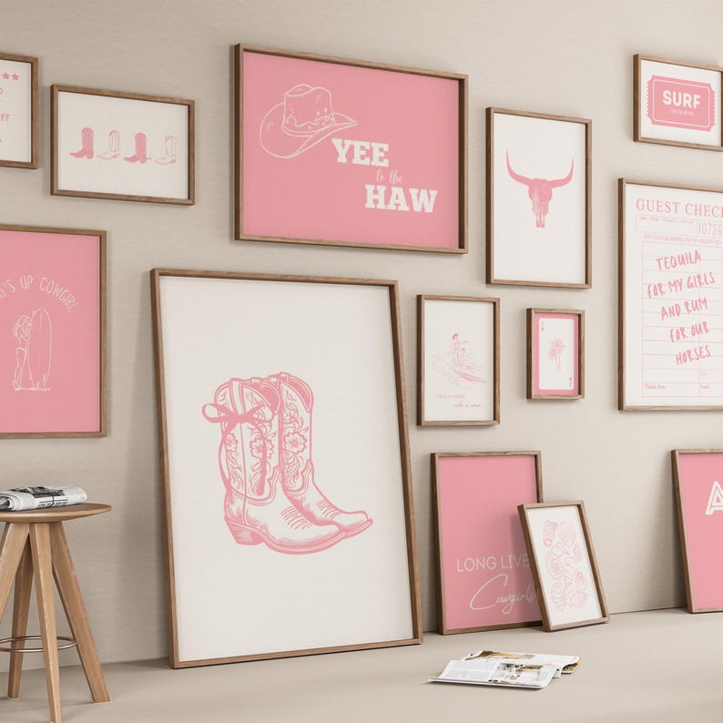 May include: A gallery wall arrangement featuring multiple framed prints with pink and white colour schemes. The artwork includes illustrations of cowboy boots, a cow skull, a surfboard, playing cards, and text such as Yee to the Haw.