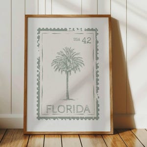 May include: Framed print featuring a vintage-style design of a palm tree, reminiscent of a postage stamp. The print includes the text "USA 42" and "FLORIDA" in a muted green color, set against a neutral background. The wooden frame adds a classic touch.
