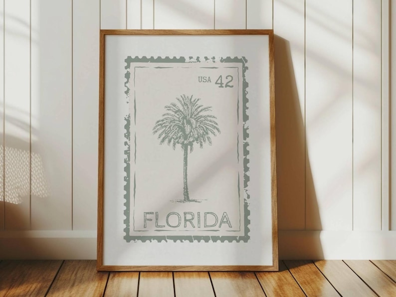 May include: Framed print featuring a vintage-style design of a palm tree, reminiscent of a postage stamp. The print includes the text "USA 42" and "FLORIDA" in a muted green color, set against a neutral background. The wooden frame adds a classic touch.