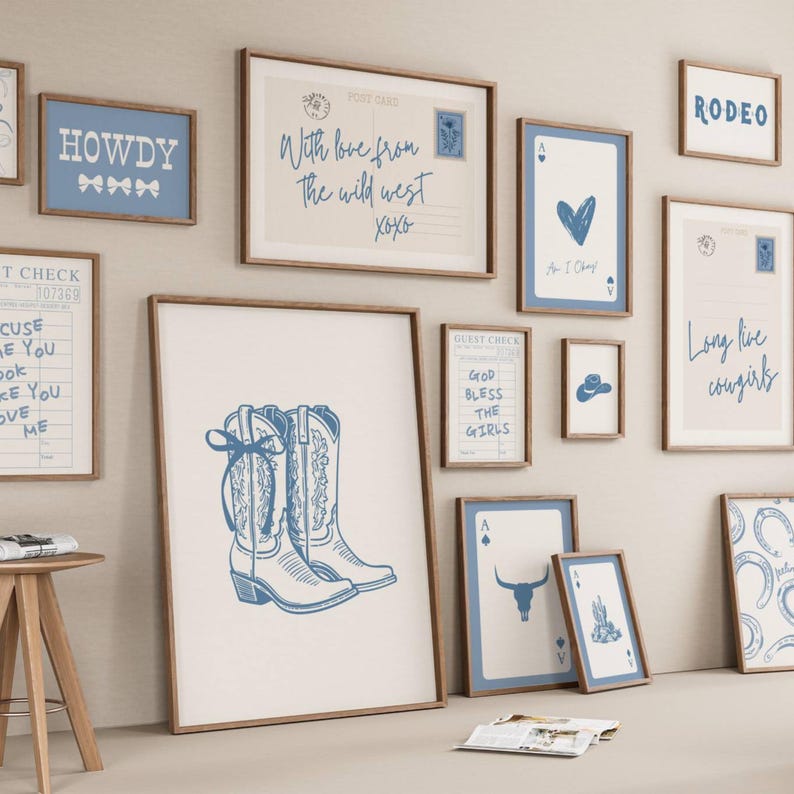 May include: A gallery wall collection of framed prints featuring blue and white western-themed illustrations, including cowboy boots, playing cards, postcards, and text like "Howdy," "Rodeo," and "Long live cowgirls" in various wooden frames.
