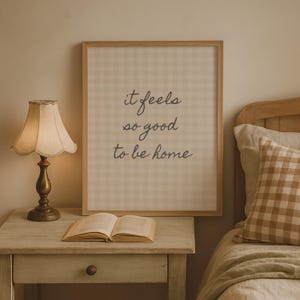 May include: A framed print with the text "it feels so good to be home" in a bedroom setting. The print has a light beige gingham pattern. A vintage-style lamp with a cream shade sits on a nightstand next to an open book and a bed with a gingham pillow.