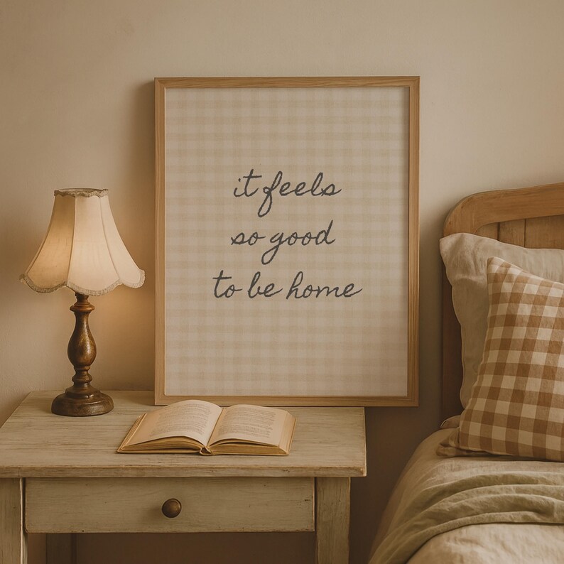 May include: A framed print with the text "it feels so good to be home" in a bedroom setting. The print has a light beige gingham pattern. A vintage-style lamp with a cream shade sits on a nightstand next to an open book and a bed with a gingham pillow.