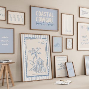 May include: A gallery wall featuring multiple framed prints with a coastal theme in shades of blue and cream. The artwork includes text such as COASTAL COWGIRL beach club, USA 42, and GUEST CHECK, alongside illustrations of palm trees, surfboards, and waves.