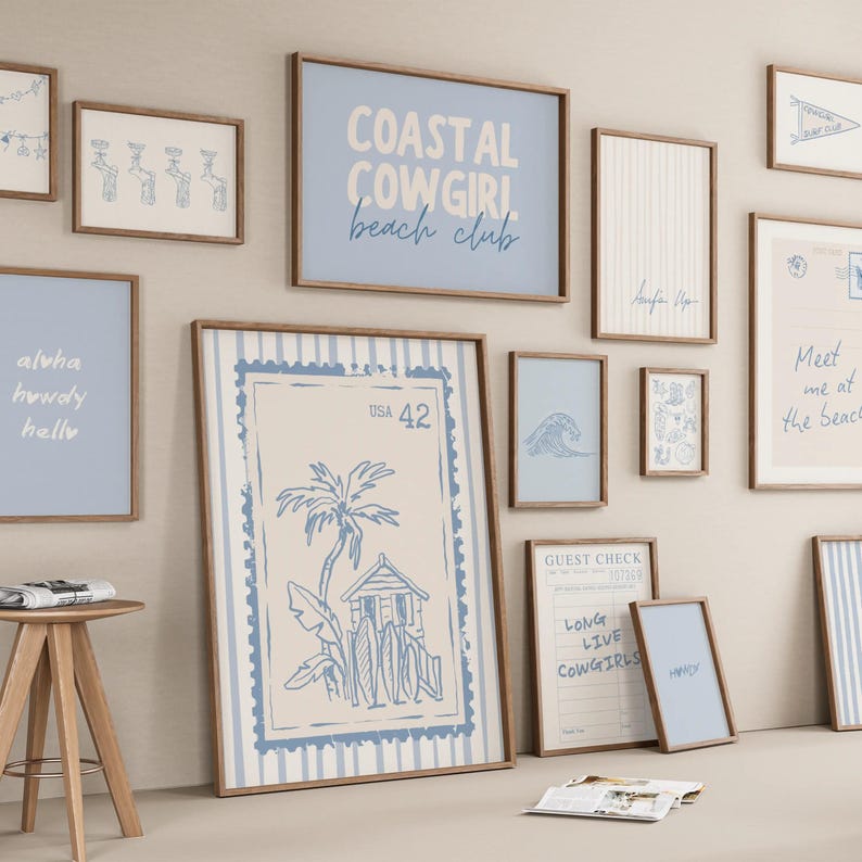 May include: A gallery wall featuring multiple framed prints with a coastal theme in shades of blue and cream. The artwork includes text such as COASTAL COWGIRL beach club, USA 42, and GUEST CHECK, alongside illustrations of palm trees, surfboards, and waves.