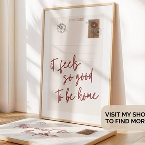 May include: Framed postcard print with the text "it feels so good to be home" in burgundy script. The print has postage stamp illustrations and a wooden frame. A second postcard is visible in the foreground.