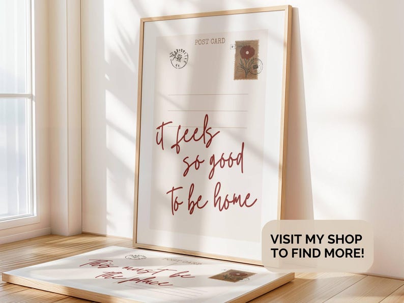 May include: Framed postcard print with the text "it feels so good to be home" in burgundy script. The print has postage stamp illustrations and a wooden frame. A second postcard is visible in the foreground.