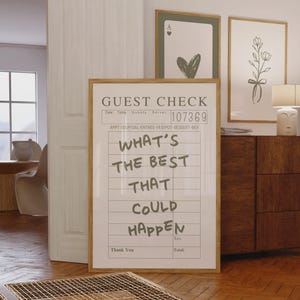 May include: A framed print designed to look like a guest check, featuring the handwritten text "WHAT'S THE BEST THAT COULD HAPPEN". The frame is light brown. Two other framed prints are visible in the background.