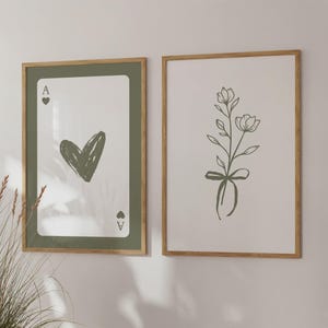 May include: Two framed art prints. One shows an Ace of Hearts playing card design with a green heart. The other print displays a floral illustration with a bow. Both are in wooden frames.