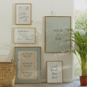 K&ouml;nnte beinhalten: Eine Sammlung gerahmter Badezimmer-Kunstdrucke mit humorvollen Zitaten und Designs. Die Drucke zeigen S&auml;tze wie "Let the good times roll" und "scrub a dub", zusammen mit Ticket-Grafiken und gestreiften Mustern. Die Rahmen sind hellbraun.