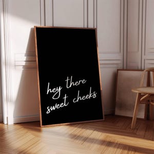 May include: A framed black art print with the phrase "hey there sweet cheeks" in white script. The frame is a warm brown color, and the print is leaning against a wall. The room has hardwood floors and a wooden chair.