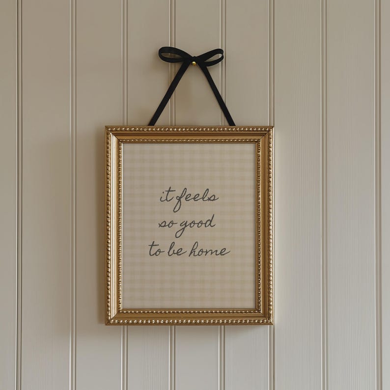 May include: A framed print with the text "it feels so good to be home." The gold-colored frame has a beaded edge and is suspended by a black ribbon tied in a bow. The background of the print has a beige and white checkered pattern.