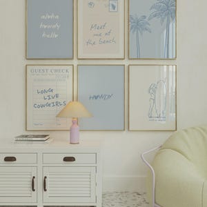 May include: A gallery wall of six framed prints in gold frames featuring blue and beige tones. The prints include text such as aloha, howdy, hello, Meet me at the beach, LONG LIVE COWGIRLS, and HOWDY, alongside illustrations of palm trees and a surfer.