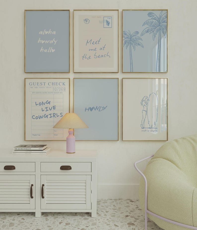 May include: A gallery wall of six framed prints in gold frames featuring blue and beige tones. The prints include text such as aloha, howdy, hello, Meet me at the beach, LONG LIVE COWGIRLS, and HOWDY, alongside illustrations of palm trees and a surfer.