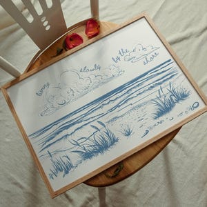May include: A framed art print featuring a blue and white illustration of a beach scene with the words "living slowly by the shore." The artwork is in a light wood frame and rests on a wooden table.