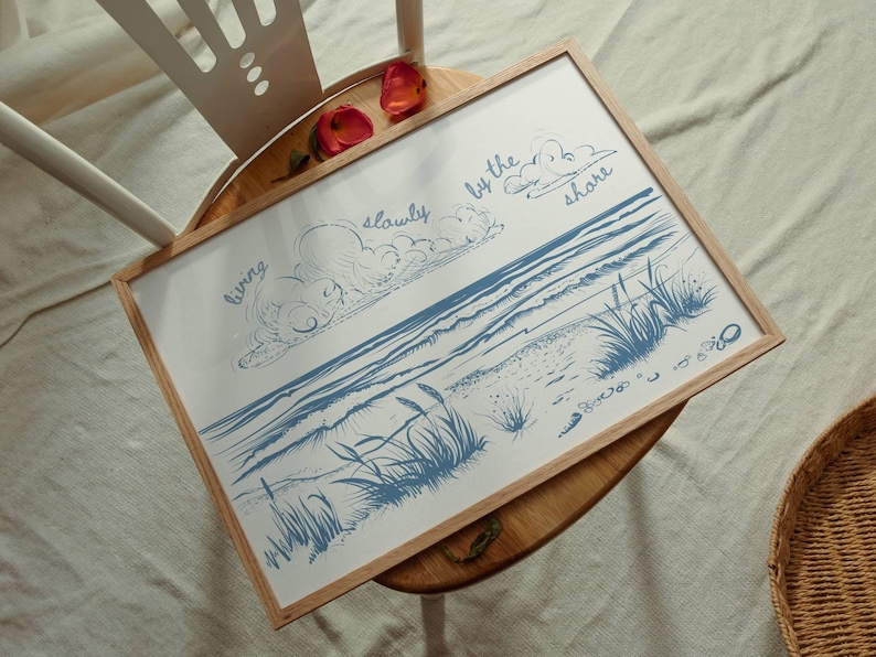 May include: A framed art print featuring a blue and white illustration of a beach scene with the words "living slowly by the shore." The artwork is in a light wood frame and rests on a wooden table.