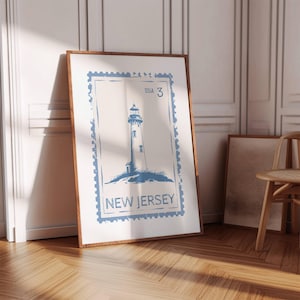 May include: A framed print featuring a blue lighthouse illustration on a white background, resembling a postage stamp. The text "USA 3" and "NEW JERSEY" are visible. The print is in a wooden frame and leans against a white wall.