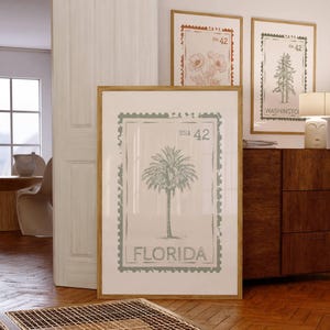May include: A framed print featuring a palm tree design with the word "FLORIDA" at the bottom. The print is in a light green color and is framed in a light brown wooden frame. Other framed prints with floral and tree designs are visible.