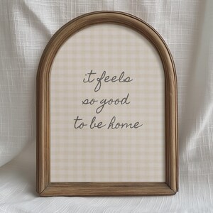 May include: Wooden framed wall art with an arched top. The artwork features a beige and white gingham pattern background with the cursive text, "it feels so good to be home."