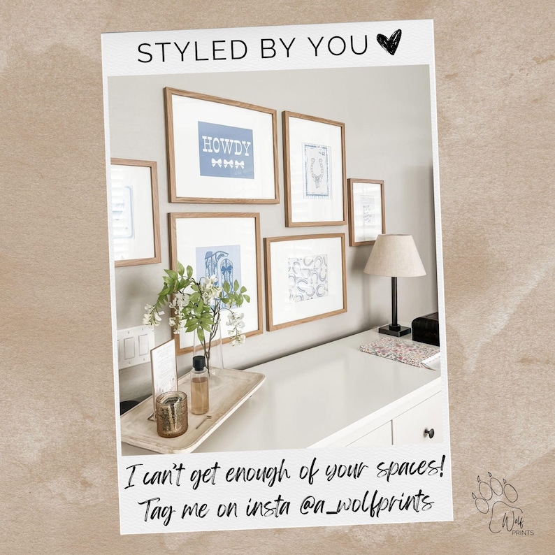 May include: A gallery wall featuring framed art prints with a neutral color palette. The prints include the word "HOWDY" and other designs in blue on white backgrounds. The wall is above a white desk with a lamp and decorative items.