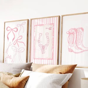 May include: Three framed art prints with a light wood finish. The prints feature pink line drawings on a white background. One print has bows, another a horseshoe, and the third a pair of cowboy boots. The print with the horseshoe has the number 42.