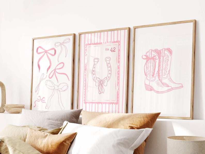 May include: Three framed art prints with a light wood finish. The prints feature pink line drawings on a white background. One print has bows, another a horseshoe, and the third a pair of cowboy boots. The print with the horseshoe has the number 42.